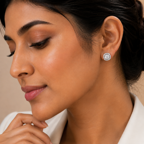 A close-up of a woman wearing a small round stud earring with a silver-toned halo setting and clear stones. The earring is shown on her ear, with her face, hand, and white collared shirt visible against a soft beige background.