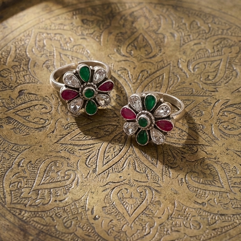 Two silver rings with floral designs set with green and magenta stones, displayed on an ornate brass plate.