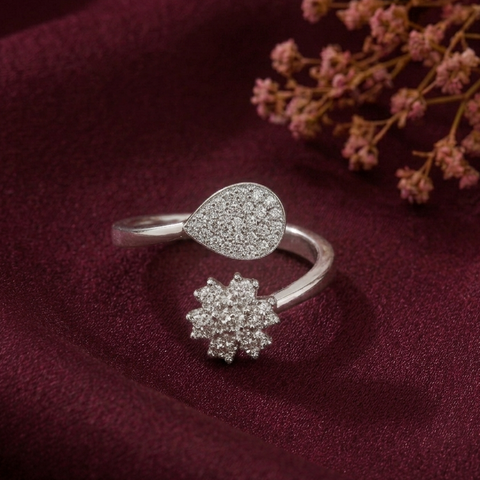A silver ring with a teardrop-shaped pavé stone top and a separate snowflake-shaped pavé stone charm, displayed on deep burgundy satin fabric with dried pink flowers in the corner.